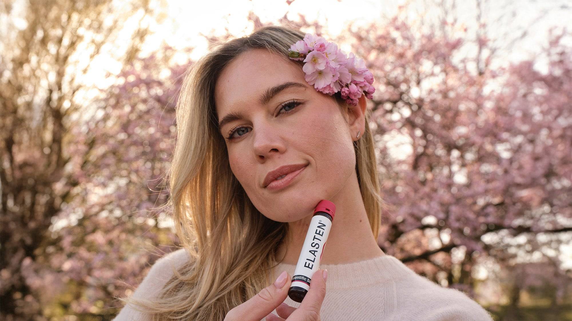 Blonde Frau hält Trinkampulle von ELASTEN® mit zwei Fingern und richtet sie in die Kamera