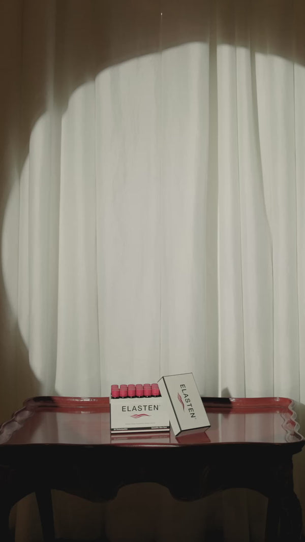 Eine dunkelhaarige Frau erscheint hinter einem Vorhang und zeigt eine ELASTEN® Trinkampulle in die Kamera