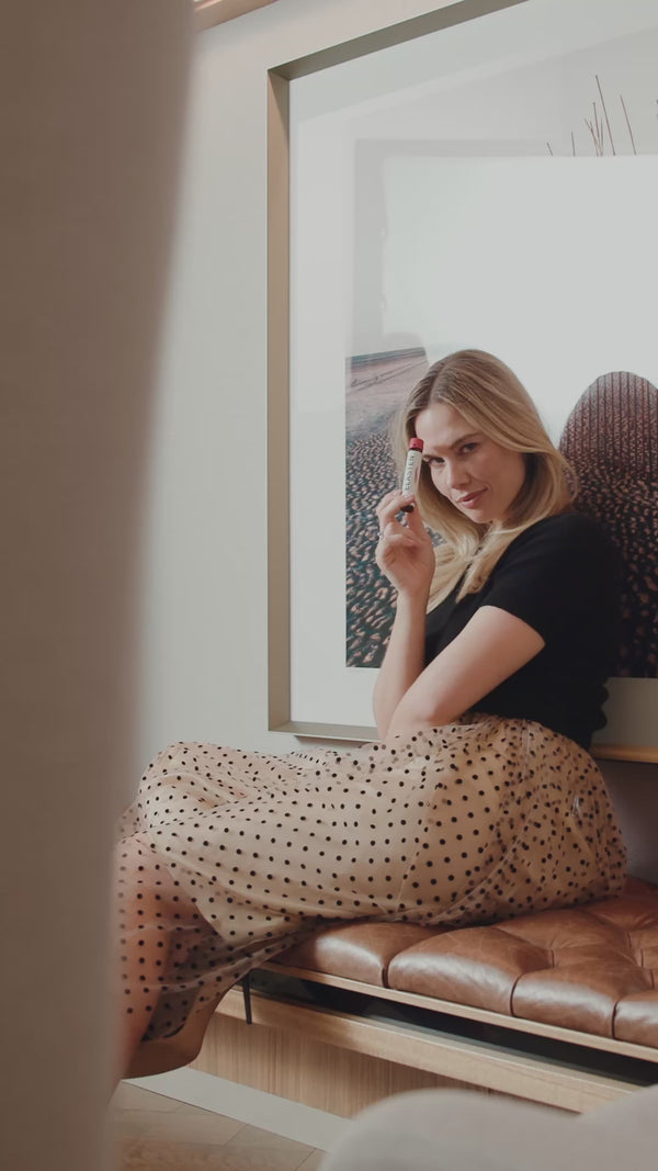 Eine blonde Frau sitzt in einer Bibliothek und zeigt eine ELASTEN® Trinkampulle in die Kamera