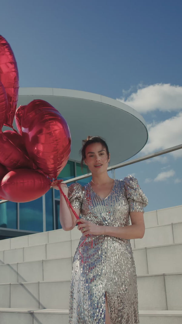 Eine dunkelhaarige Frau hält Ballons in Herzform in der einen Hand und zeigt eine ELASTEN® Trinkampulle mit der anderen Hand in die Kamera