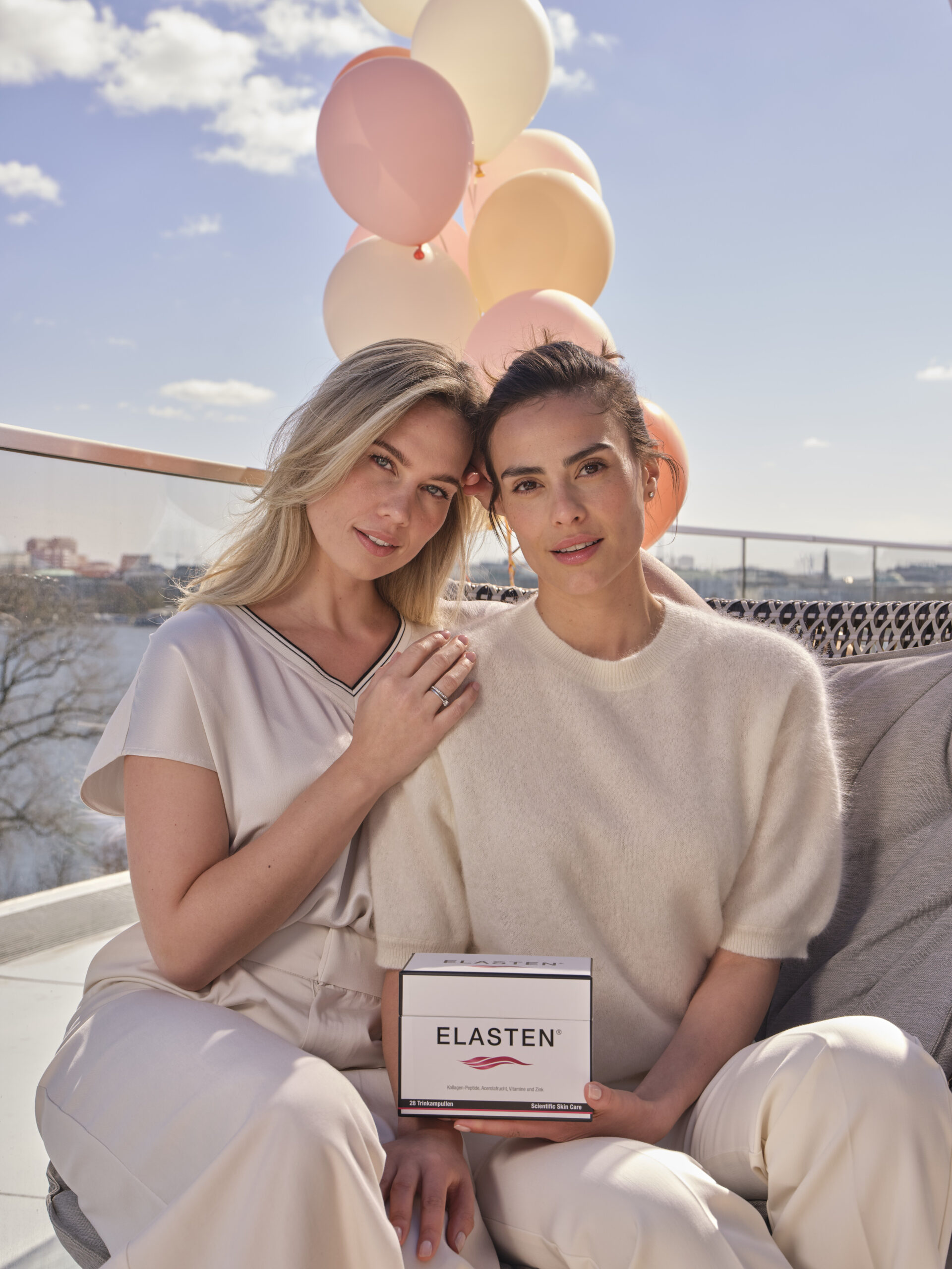 Auf dem Bild sind zwei Frauen Mitte 30 zu sehen, von denen eine blond und eine brünett ist, tragen beide helle Kleidung und sie sitzen nebeneinander auf einem Sessel an der frischen Luft, halten eine Packung ELASTEN in der Hand und im Hintergrund sieht man helle Luftballons vor blauem Himmel.